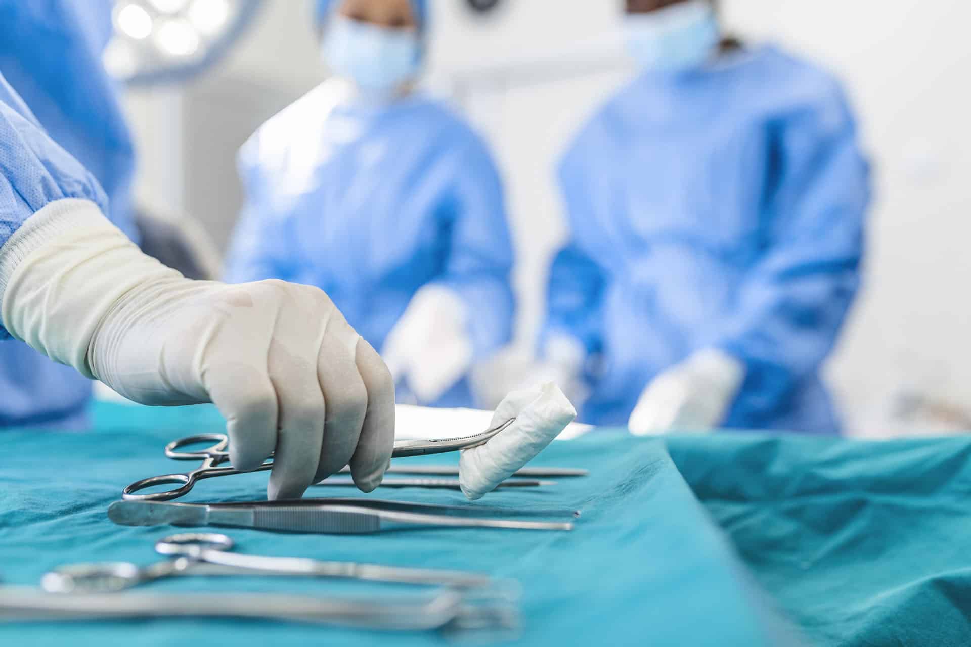 Nurse hand taking surgical instrument for group of surgeons at background operating patient in surgical theatre. Steel medical instruments ready to be used. Surgery and emergency concept